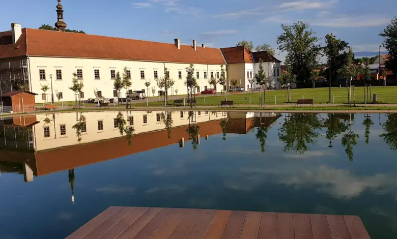 Velká budova se zrcadlí v klidné vodě s lavičkami.