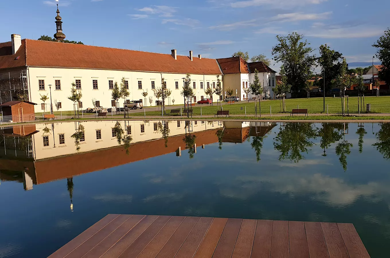 Velká budova se zrcadlí v klidné vodě s lavičkami.