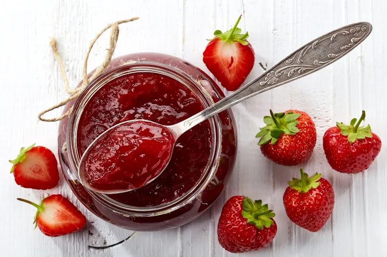 Sklenice domácí jahodové marmelády s lžící a čerstvými jahody.