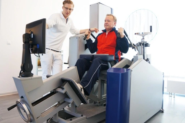Muž cvičí na rehabilitačním stroji pod dohledem fyzioterapeuta.