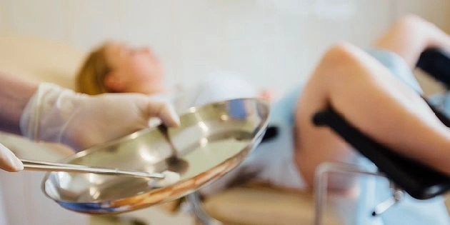 Ruce s rukavicemi drží nástroje pro kyretáž u pacientky na vyšetřovacím stole.