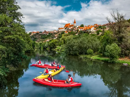 Lidé plavou na kajakech řekou směrem k vesnici.